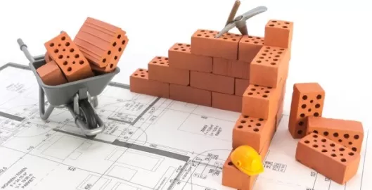 Baustelle mit Mauer aus roten Ziegelsteinen, Schubkarre mit Ziegeln und gelbem Bauhelm