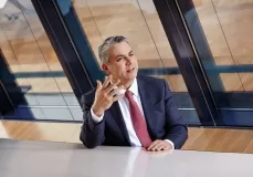 Mann in dunklem Anzug und roter Krawatte sitzt an einem Tisch vor großen Fenstern und gestikuliert mit der rechten Hand