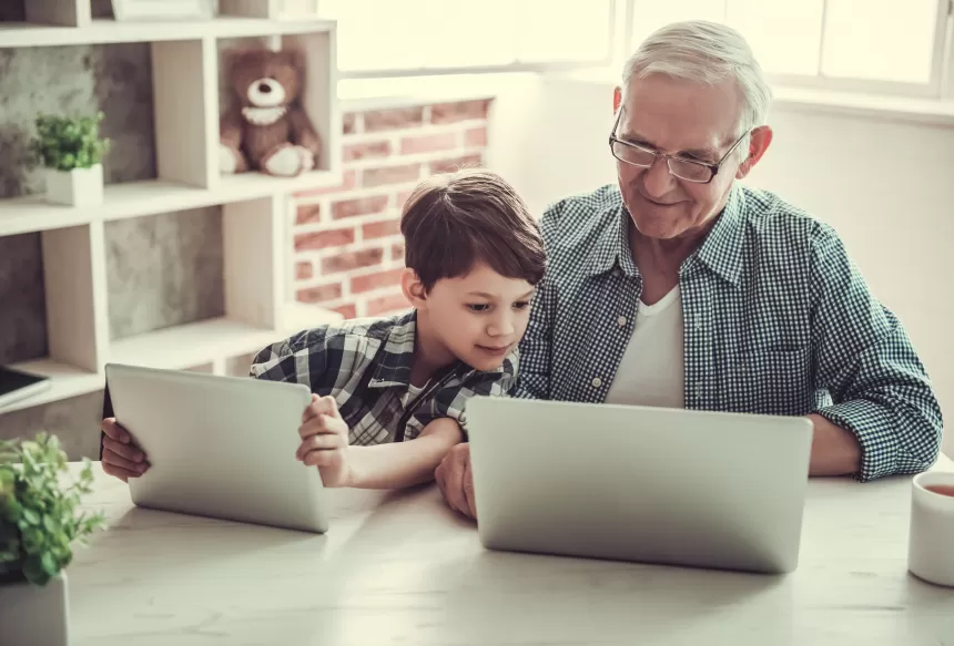 Ein Mann und ein Junge sitzen an einem Tisch mit vor ihnen geöffneten Laptops, drinnen. Die Umgebung umfasst Möbel und eine Wand im Hintergrund.
