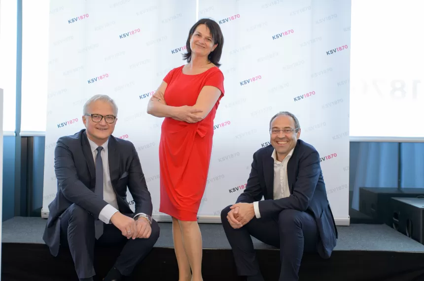 Frau in rotem Kleid steht mit verschränkten Armen auf einer Bühne, zwei Männer in Anzügen sitzen neben ihr auf der Bühne