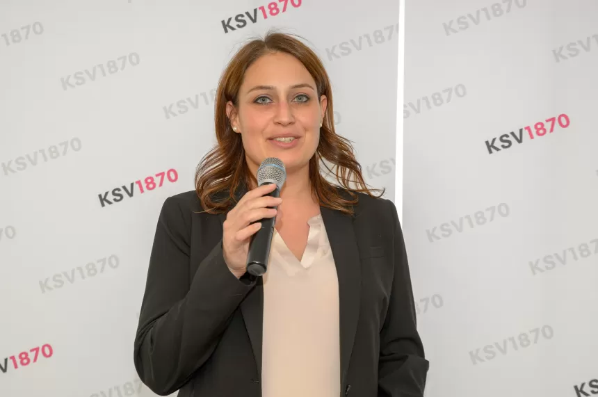 Frau mit braunen, schulterlangen Haaren hält Mikrofon, trägt schwarzen Blazer und helles Oberteil, steht vor KSV1870-Hintergrund