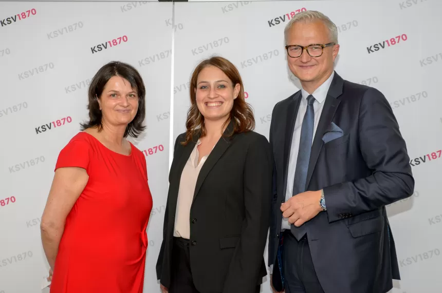 Drei Personen stehen nebeneinander vor einer Wand mit 'KSV1870'-Logo, zwei Frauen und ein Mann in formeller Kleidung