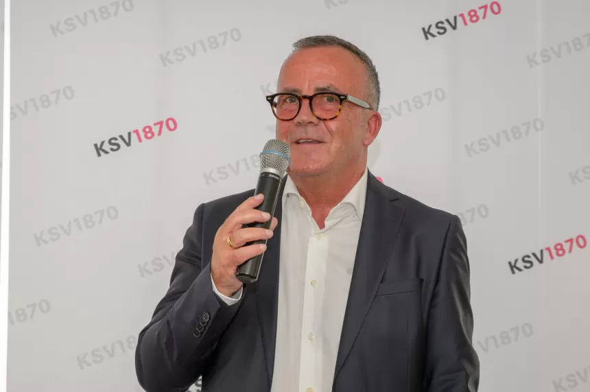 Mann in dunklem Anzug und weißem Hemd hält ein Mikrofon vor einem Hintergrund mit dem Schriftzug 'KSV1870'