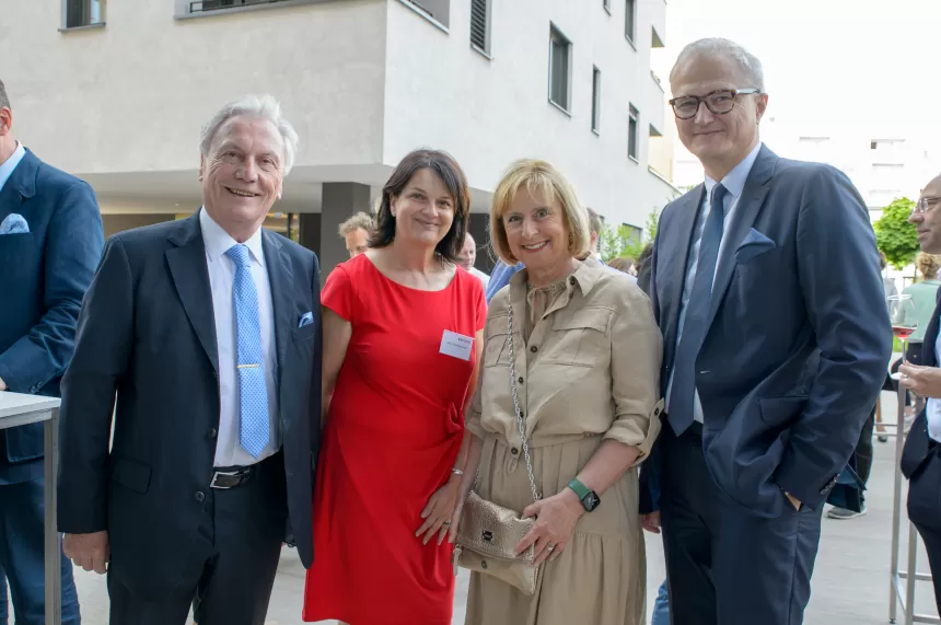 Vier Personen stehen vor einem modernen Gebäude, dem neuen KSV1870 Standort in Linz, zwei Männer in Anzügen, eine Frau in rotem Kleid, eine Frau in beigem Kleid mit Tasche