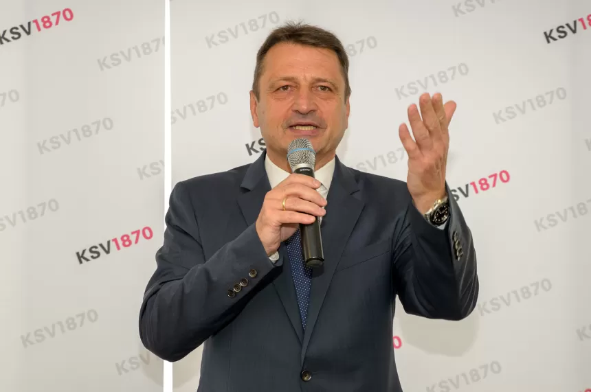 Mann in dunklem Anzug hält Mikrofon und gestikuliert mit der rechten Hand vor einer Wand mit KSV1870-Logo