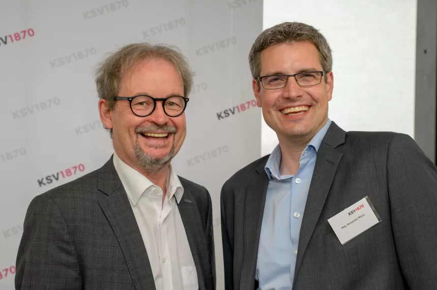 Zwei Männer, Hannes Frech und Alexander Mitter, in Anzügen mit Hemden stehen nebeneinander vor einer Wand mit KSV1870-Logo
