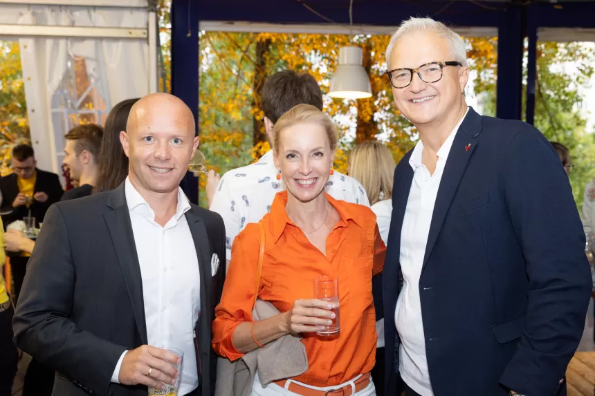 Drei Personen beim Sommerausklang des KSV1870, zwei Männer in Anzügen und eine Frau in orangefarbener Bluse, halten Getränke in der Hand