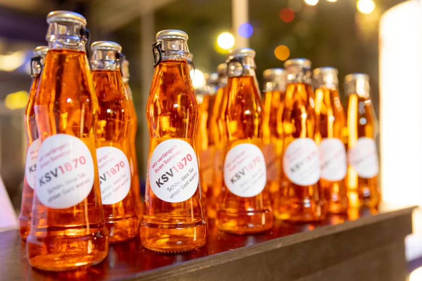 Viele kleine orange Getränkeflaschen sind hintereinander aufgereiht und haben einen Sticker mit der Aufschrift 'Wir verlängern Ihren Sommer. KSV1870 - Schön, dass Sie unser Gast waren'