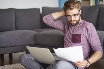 Mann mit Brille sitzt auf dem Boden vor einem Sofa, liest konzentriert ein Dokument und hat einen Laptop auf dem Schoß