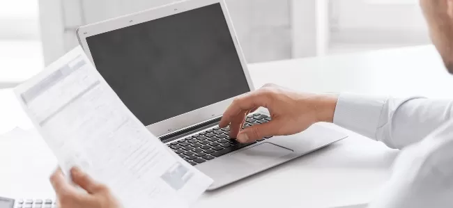 Person hält Vertragspapier vor Laptop und zeigt mit Stift darauf