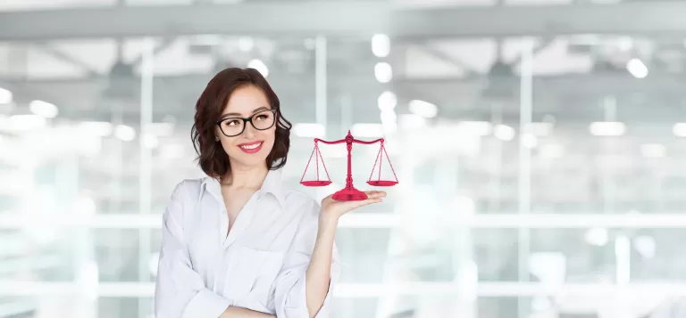 Frau mit Brille hält eine rote Waage der Gerechtigkeit in der Hand vor einem hellen Bürohintergrund