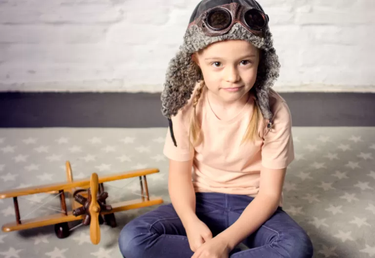 Kind sitzt auf Sternenmuster-Teppich, trägt Fliegerkappe mit Brille, neben ihm ein hölzernes Spielzeugflugzeug, symbolisch für Jungunternehmer