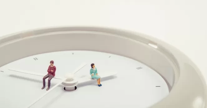 Zwei kleine Figuren sitzen auf den Zeigern einer großen Uhr, eine Person in rotem Oberteil und eine in grünem Kleid.