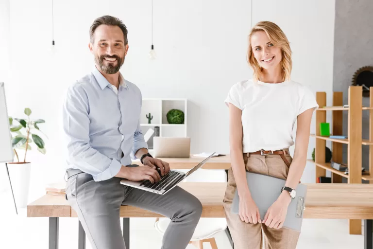 Zwei junge Erwachsene in einem modernen Büro, eine Person sitzt mit Laptop auf einem Tisch, die andere steht daneben mit einem Ordner in der Hand