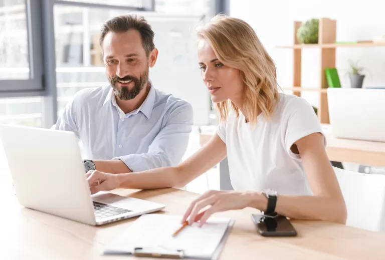 Zwei Personen sitzen an einem Tisch mit Laptop und Notizblock, arbeiten gemeinsam in einem hellen Büro