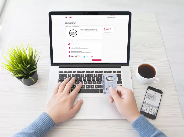 Person hält Personalausweis vor Laptop mit InfoPass-Seite, daneben Smartphone und Kaffeetasse auf weißem Tisch