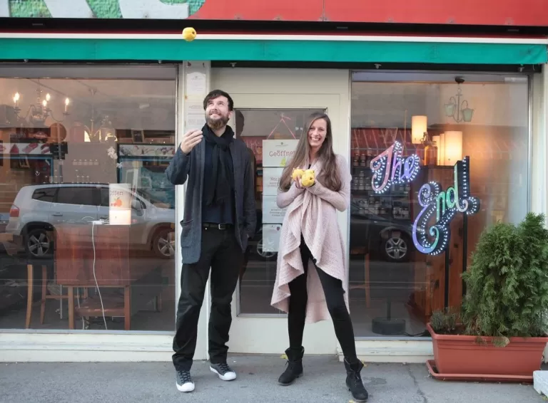 Ein Mann und eine Frau stehen lächelnd vor einem Geschäft mit Äpfeln in der Hand. Die Szene spielt sich im Freien auf einer Straße ab.