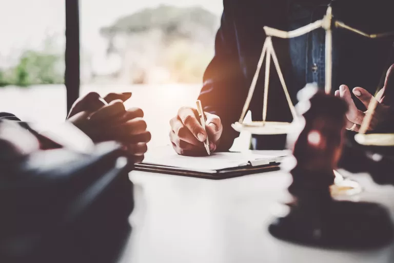 Person unterschreibt Dokument am Tisch mit Justizwaage und Hammer im Vordergrund