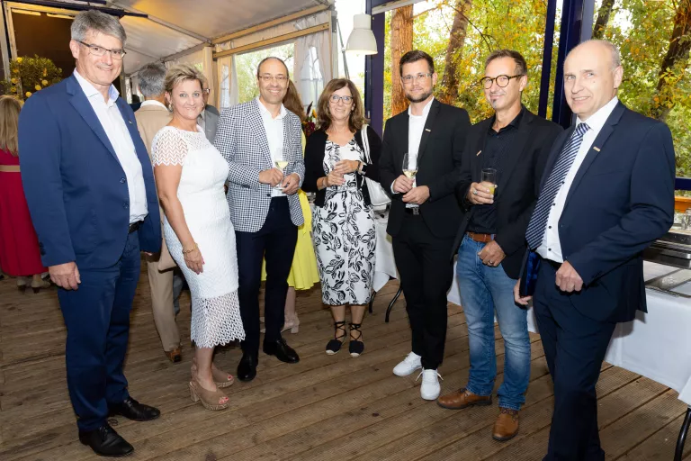 Sieben Personen in formeller Kleidung stehen in einem Raum mit großen Fenstern und Holzboden, einige halten Getränke in der Hand.