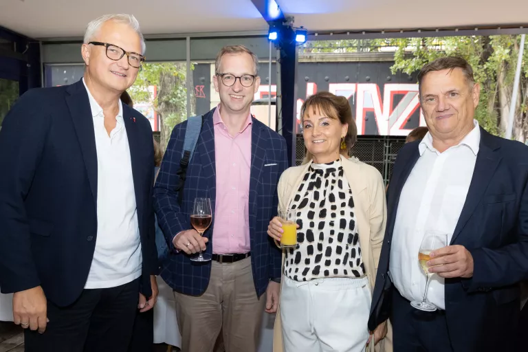 Vier Personen beim Sommerausklang des KSV1870, zwei Männer tragen Anzüge, eine Frau trägt weiße Hose und gemustertes Oberteil, alle halten Getränke