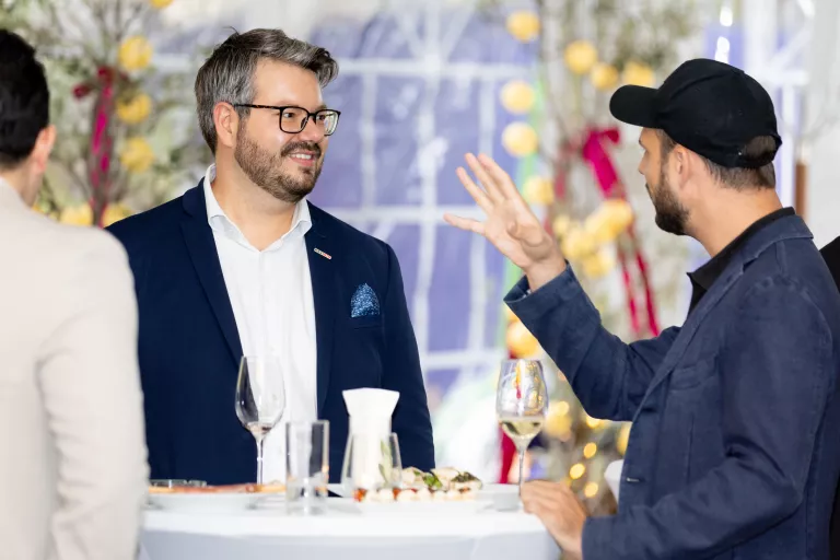 Zwei Männer im Gespräch am Stehtisch beim Sommerausklang des KSV1870, einer trägt Anzug mit weißem Hemd, der andere dunkle Jacke und schwarze Kappe