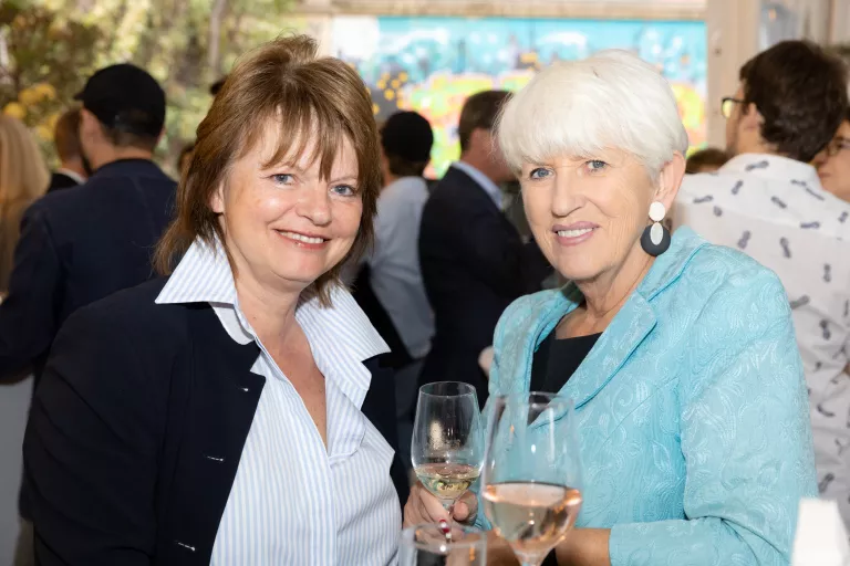 Zwei Frauen beim Sommerausklang des KSV1870, eine trägt weiße Bluse und dunkle Jacke, die andere hellblaue Jacke, beide halten Weingläser