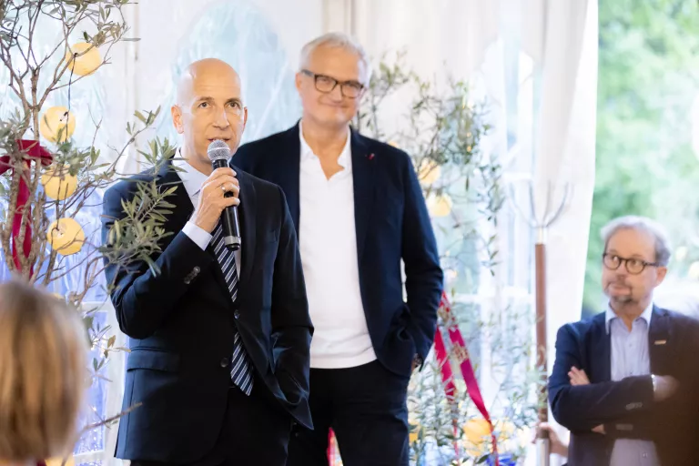 Zwei Männer in Anzügen sprechen beim Sommerausklang des KSV1870, einer hält ein Mikrofon, im Hintergrund Pflanzen und weiße Vorhänge