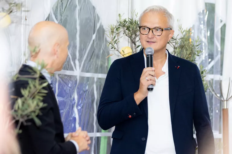 Zwei Männer in Anzügen sprechen beim Sommerausklang des KSV1870, einer hält ein Mikrofon, im Hintergrund Pflanzen und weiße Vorhänge