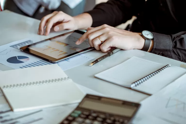 Eine Person sitzt an einem Schreibtisch mit Zetteln, Blöcken, einem Taschenrechner und sieht sich auf einem Tablett verschiedene Diagramme an