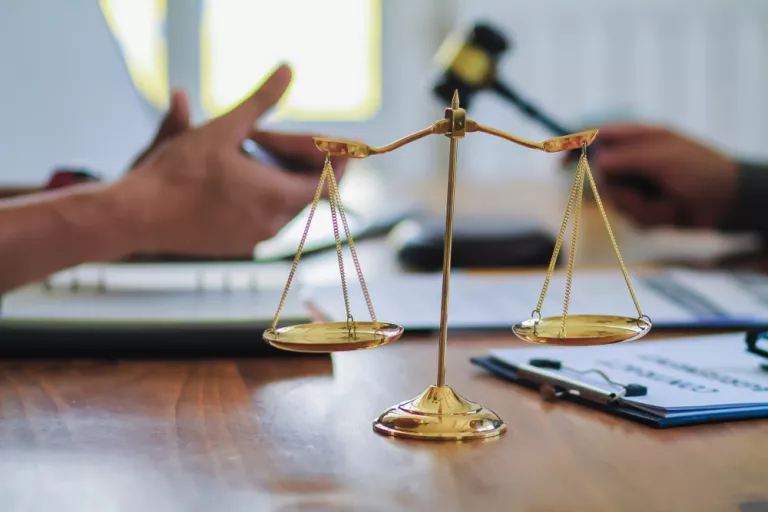 Eine Justizwaage im Vordergrund, die Hände zweier Menschen im Hintergrund, einer davon hält einen Justizhammer