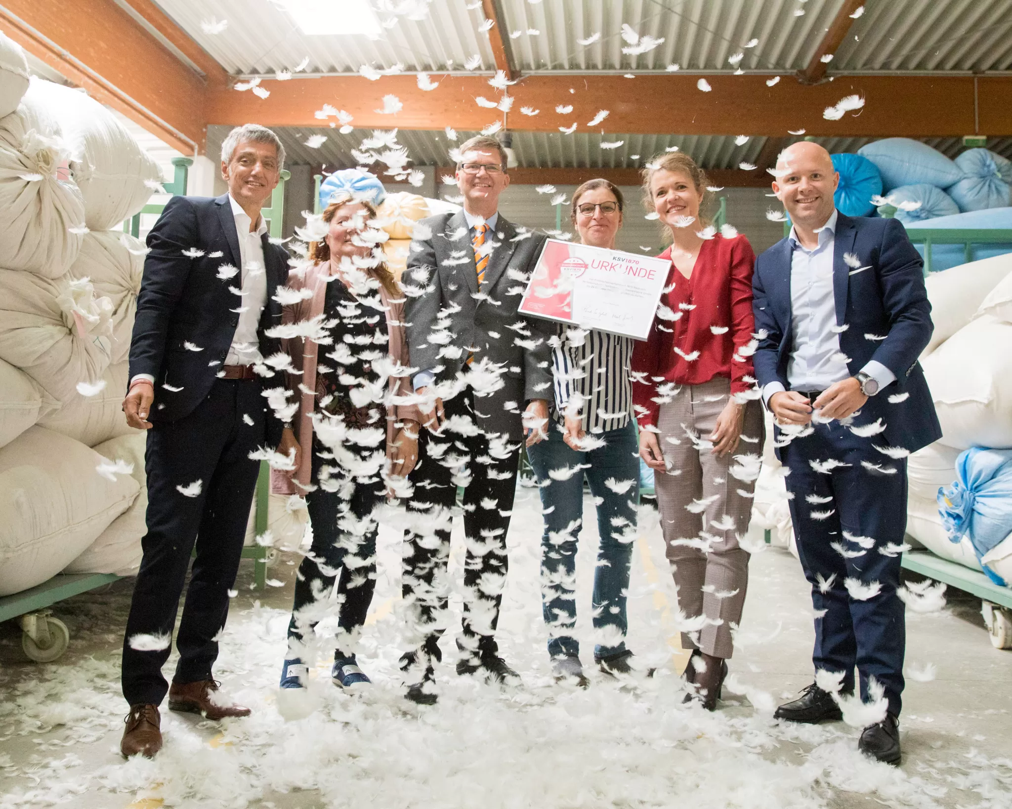 6 Personen stehen in einem Matratzenlager, eine Person hält eine Urkunde. Im Vordergrund fliegen Kissenfedern