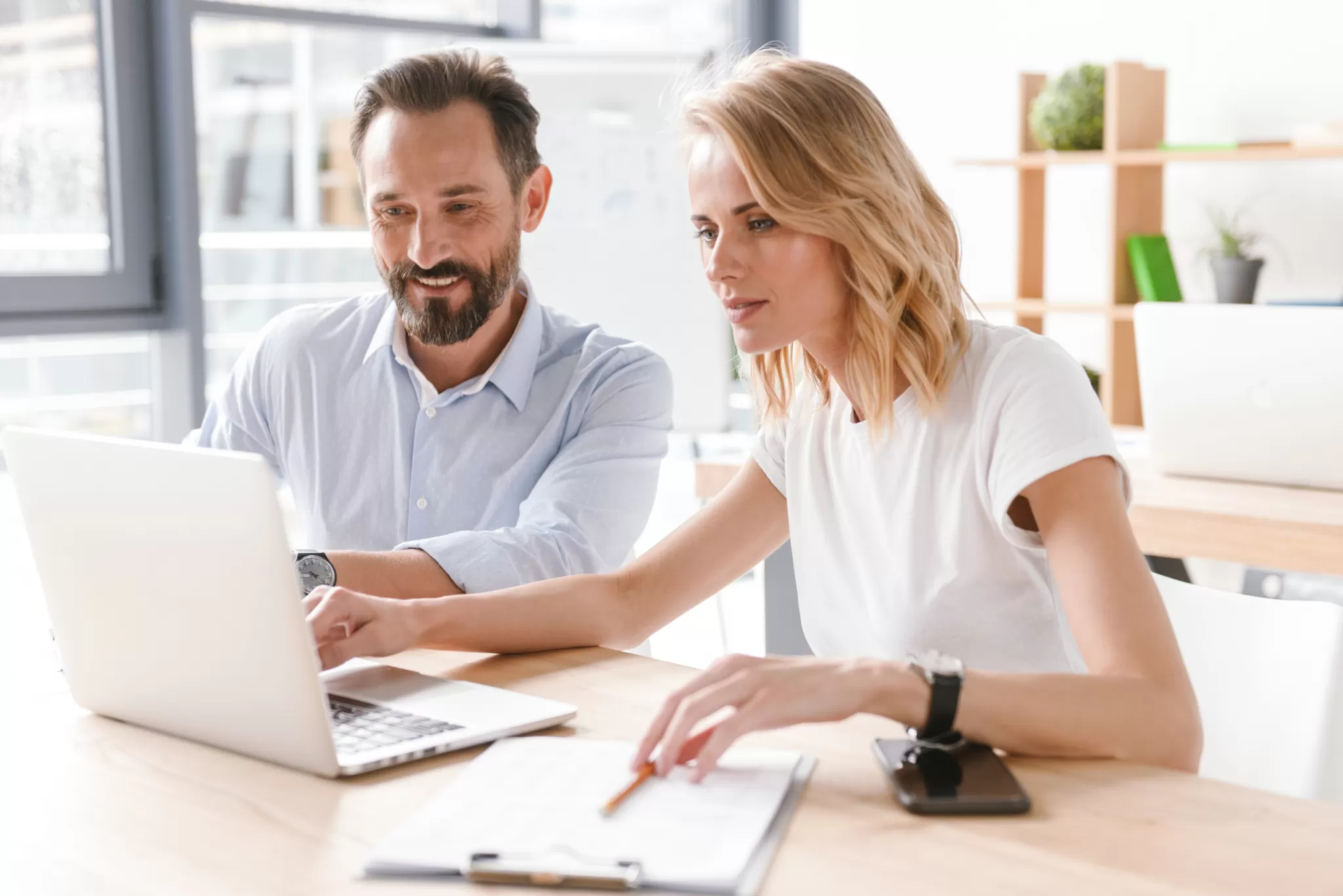 Zwei Personen sitzen an einem Tisch mit Laptop und Notizblock, arbeiten gemeinsam in einem hellen Büro
